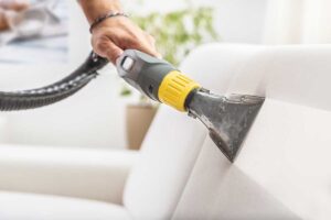 A person vacuuming a living room while sunlight streams through large windows onto tidy furniture.