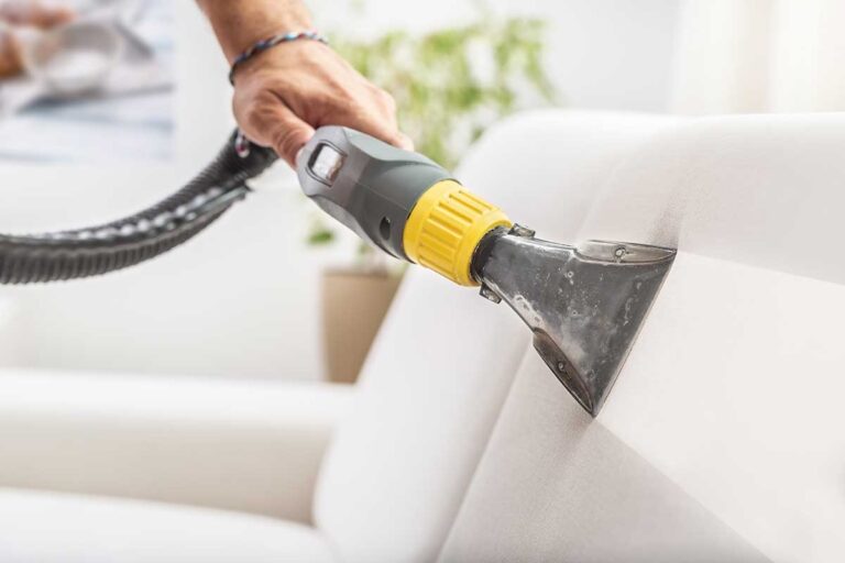 A person vacuuming a living room while sunlight streams through large windows onto tidy furniture.