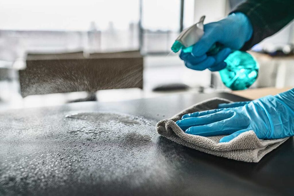 Person dusting a living room shelf with a microfiber cloth, bucket of cleaning supplies visible nearby.