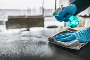 Person dusting a living room shelf with a microfiber cloth, bucket of cleaning supplies visible nearby.
