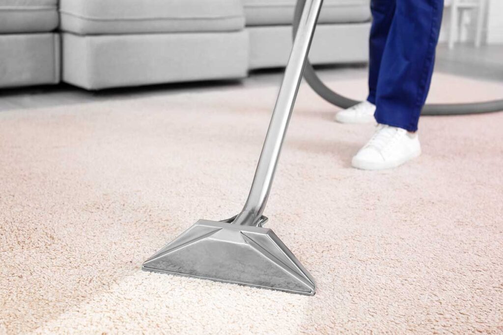 Person vacuuming a bright living room beside organized cleaning supplies and a basket of folded linens.