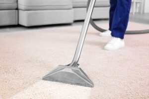 Person vacuuming a bright living room beside organized cleaning supplies and a basket of folded linens.
