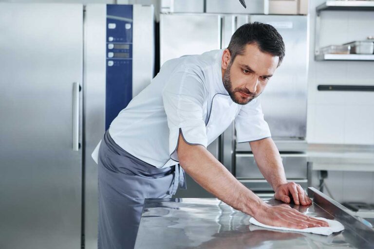 Person wiping a stainless refrigerator beside clean shelves, drawers, and kitchen cleaning cloths.